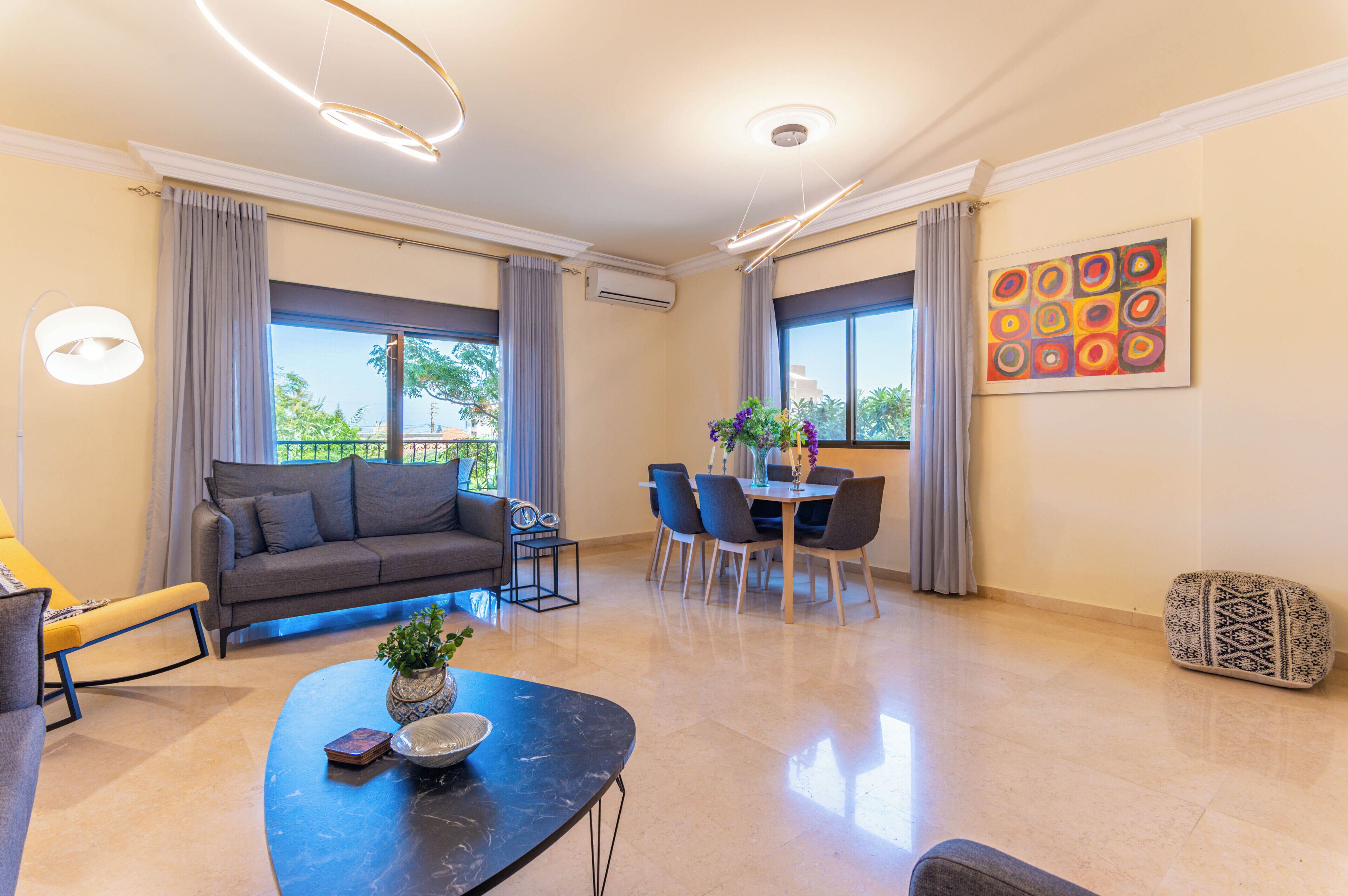 Una sala de estar moderna con sofá, mesa de comedor y sillas, ventanas amplias con cortinas y una lámpara de techo.