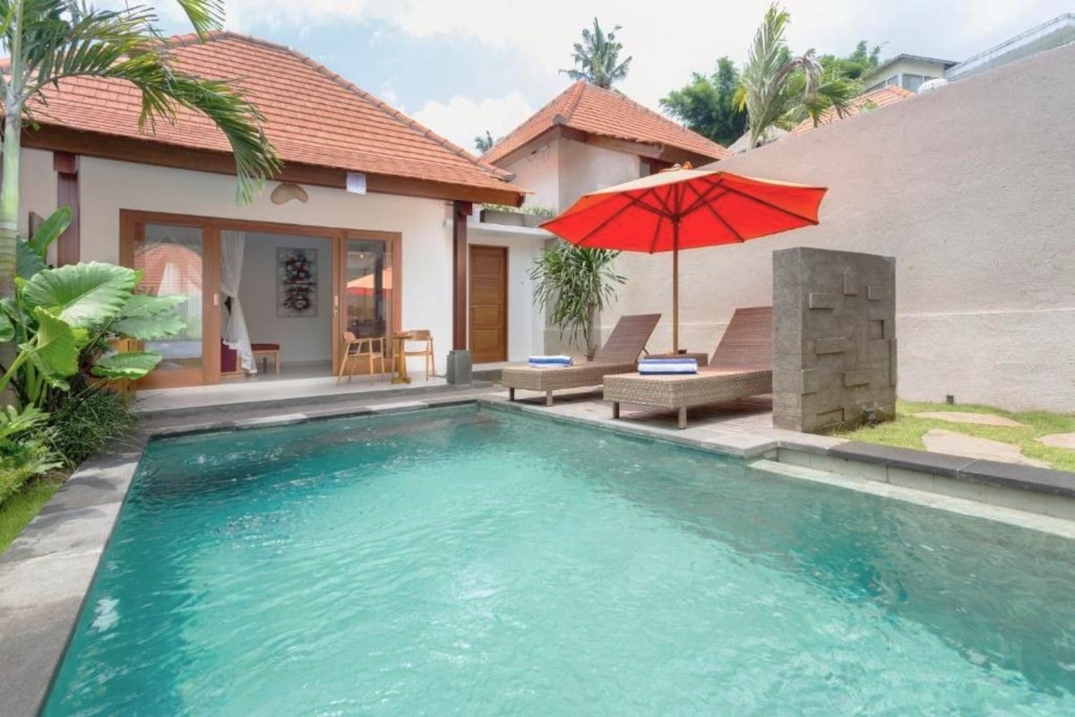 Una piscina con sillones de descanso y una sombrilla roja, rodeada por un edificio con tejado de baldosas y un muro de piedra.