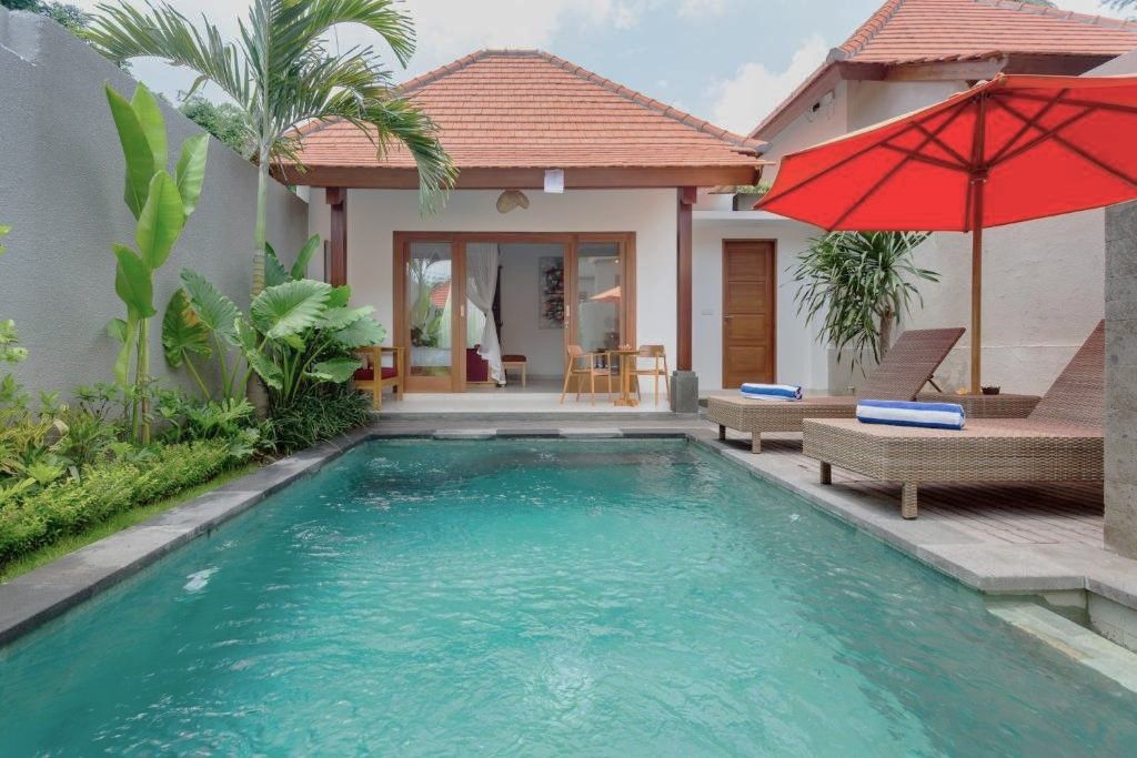 A swimming pool with lounge chairs and a red umbrella, surrounded by greenery and a building with a tiled roof.