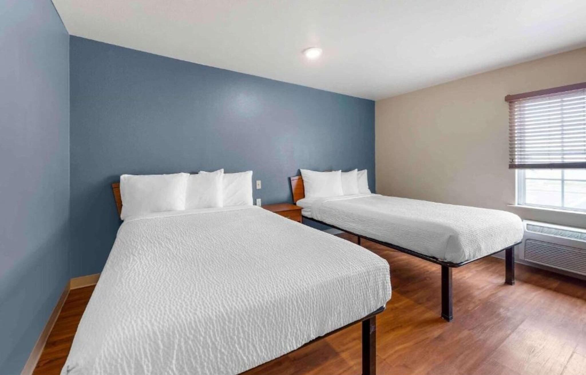 Une chambre avec deux lits simples, un plancher en bois, un mur d’accent bleu et une fenêtre avec des stores.