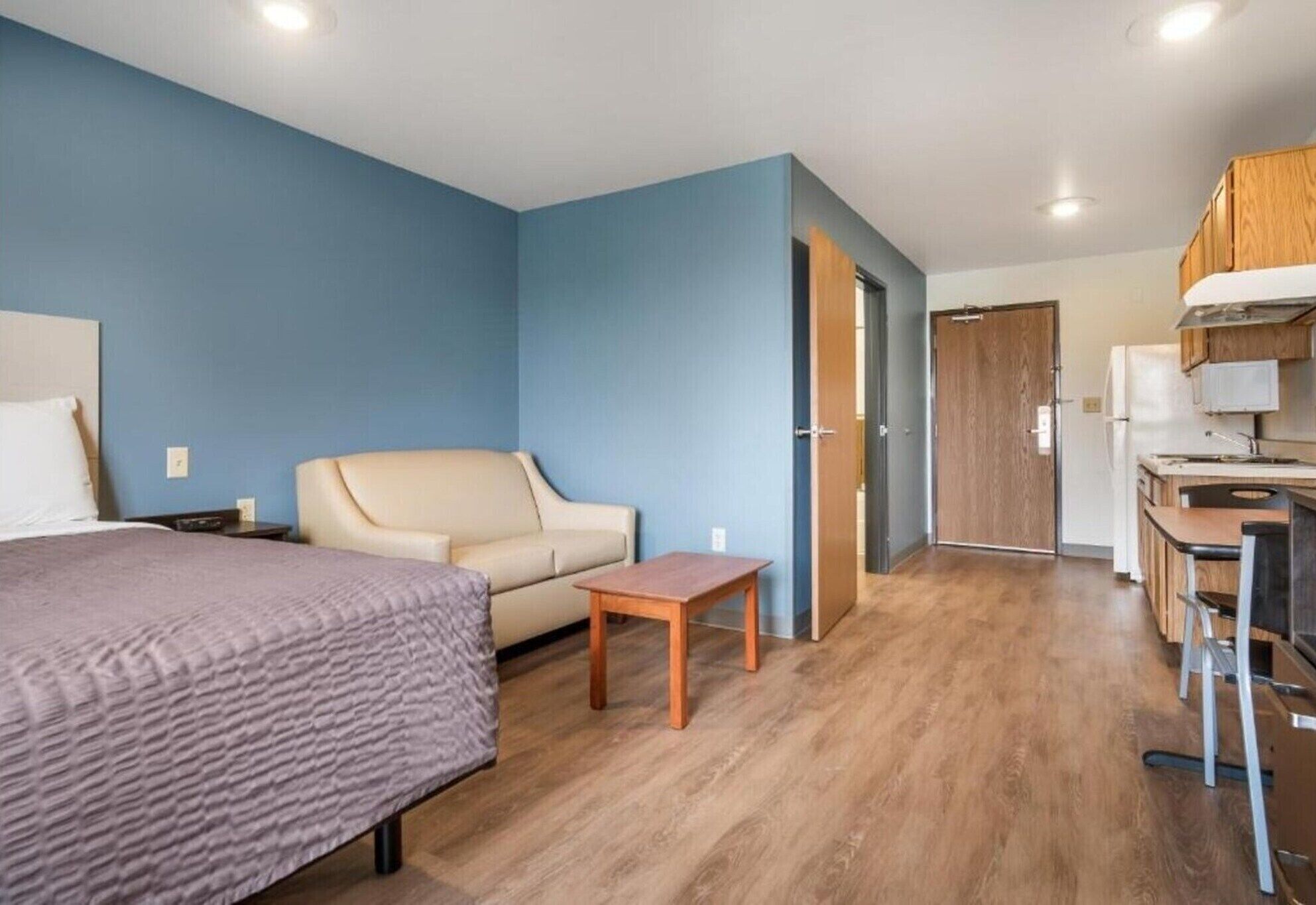 Une chambre à coucher comprenant un lit, un canapé, une petite table, un coin cuisine avec un réfrigérateur et une porte en bois.