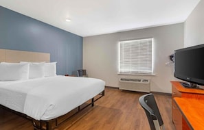 Quarto de hotel com cama, escrivaninha, cadeira, televisão e janela com persianas.