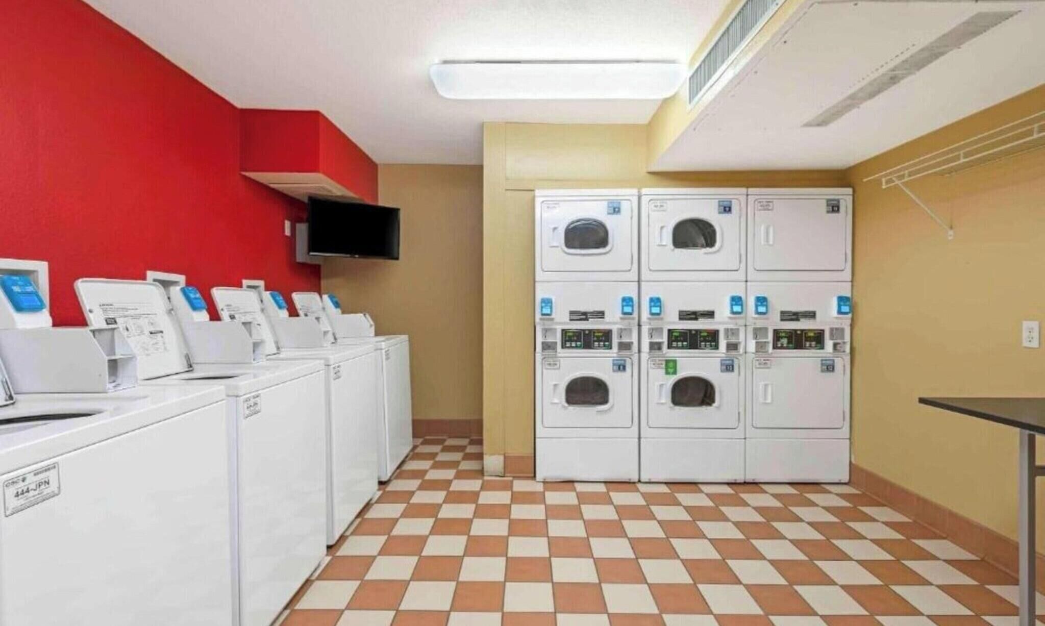Uma lavanderia com várias máquinas de lavar e secadoras, uma televisão de tela plana fixada na parede e um padrão de piso quadriculado.