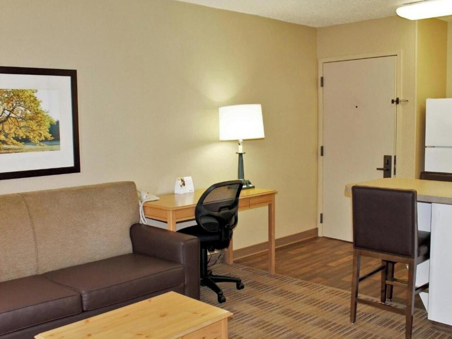 Quarto de hotel com sofá de couro marrom, uma escrivaninha de madeira com luminária, um computador e uma cadeira.