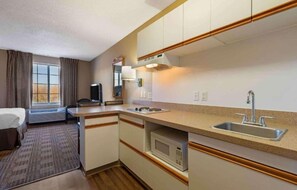 Ett kompakt hotellrum med ett litet kök, en säng, en TV och ett fönster med gardiner.