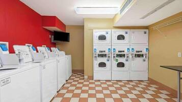 Une buanderie équipée de plusieurs machines à laver et de plusieurs sèche-linge, un téléviseur à écran plat fixé au mur et un carrelage à motifs à carreaux.
