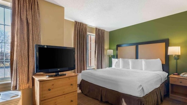 Habitación de hotel con cama, televisor sobre un aparador de madera, mesita de noche con lámpara y vista al exterior.