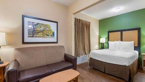 Una habitación de hotel con cama, sofá, una mesita, un cuadro en la pared y una ventana con cortinas.