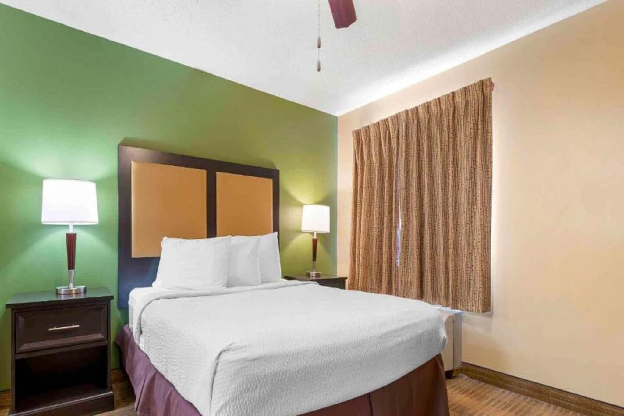 Une chambre d’hôtel avec un lit, deux tables de chevet avec des lampes, une tête de lit et une fenêtre avec des rideaux.