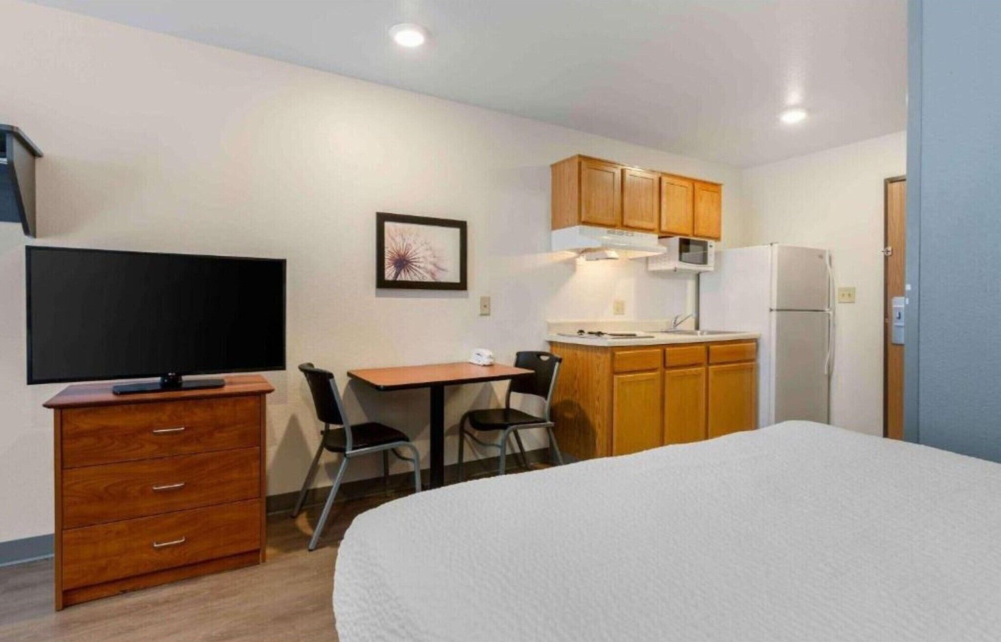 A compact hotel room with a bed, a TV, a desk with chairs, a kitchenette, and wooden cabinets.