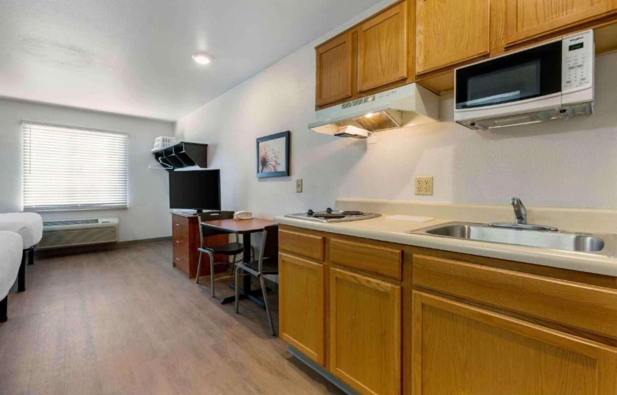 A compact kitchen with wooden cabinets, a microwave, a sink, and a small dining area with a chair and table.