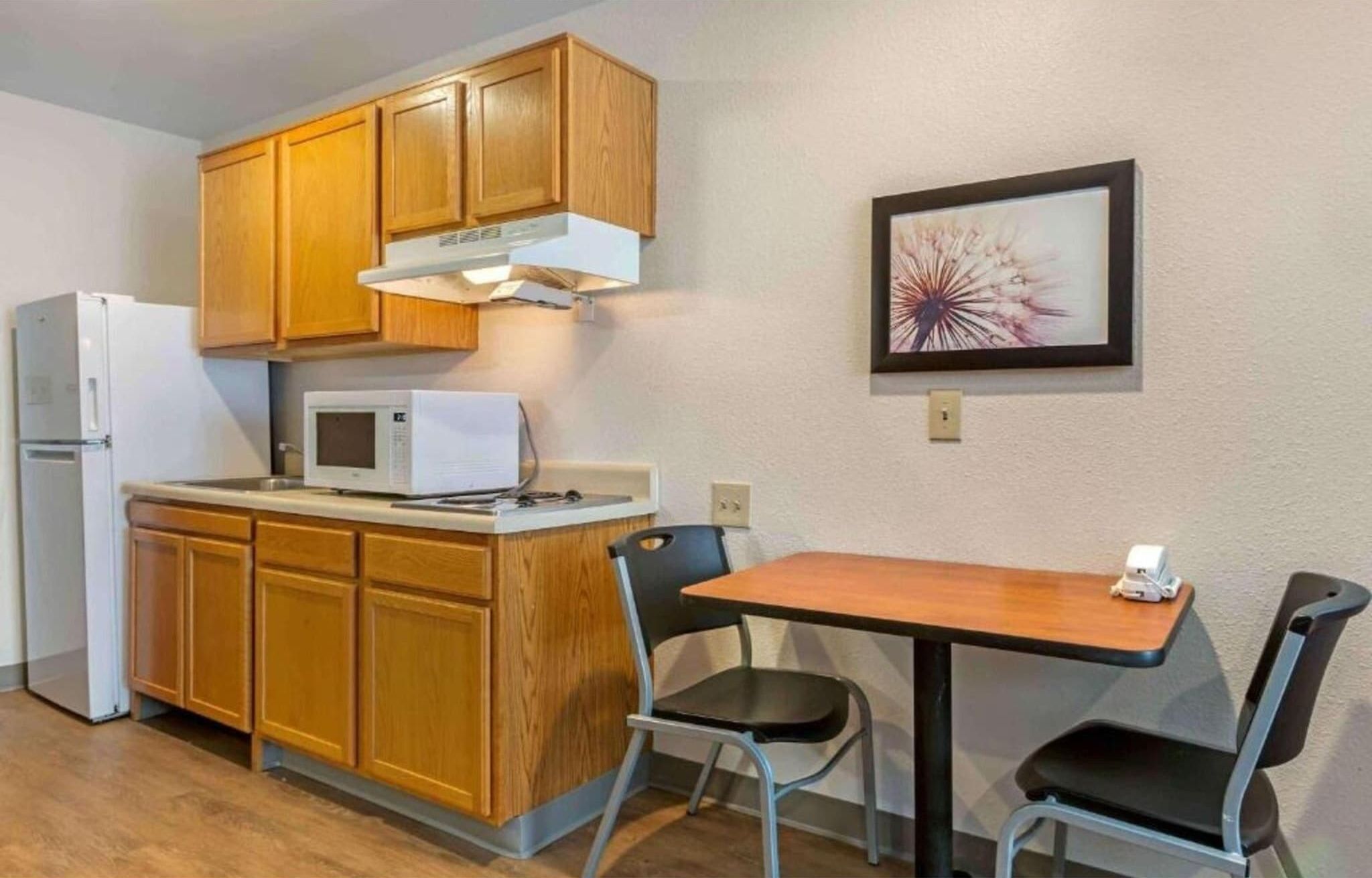 Cocina compacta con armarios de madera, microondas, refrigerador y una pequeña mesa de comedor con dos sillas.