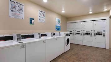 Une buanderie équipée de plusieurs machines à laver et de plusieurs sèche-linge, avec un sol carrelé et des affiches informatives accrochées aux murs.