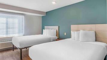 A hotel room with two beds, a window with blinds, a wall heater, and a ceiling light.
