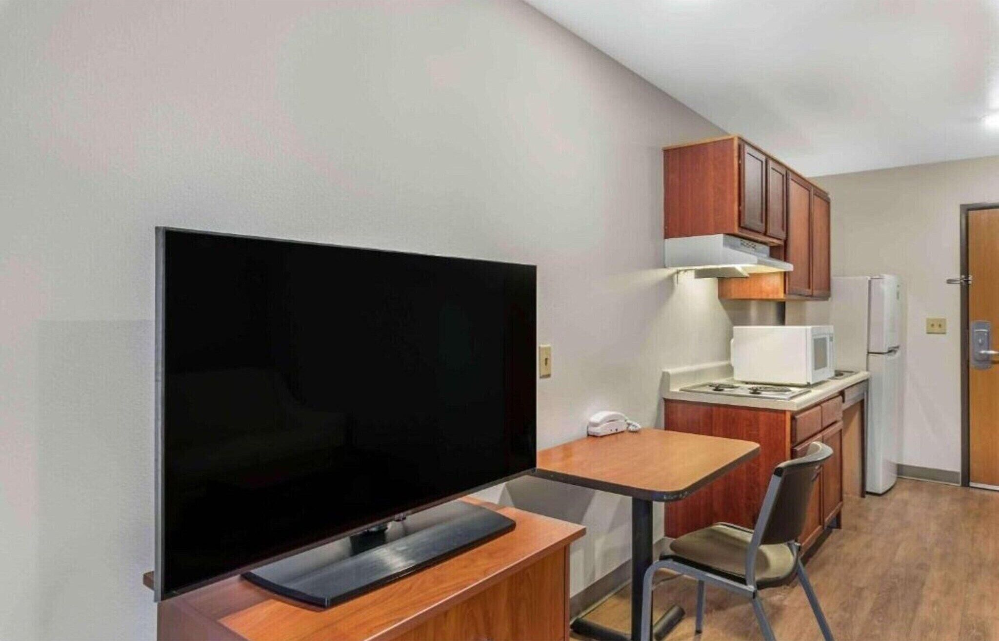 A compact kitchenette with a microwave, stove, and cabinets, a small dining table with a chair, and a TV on a stand.
