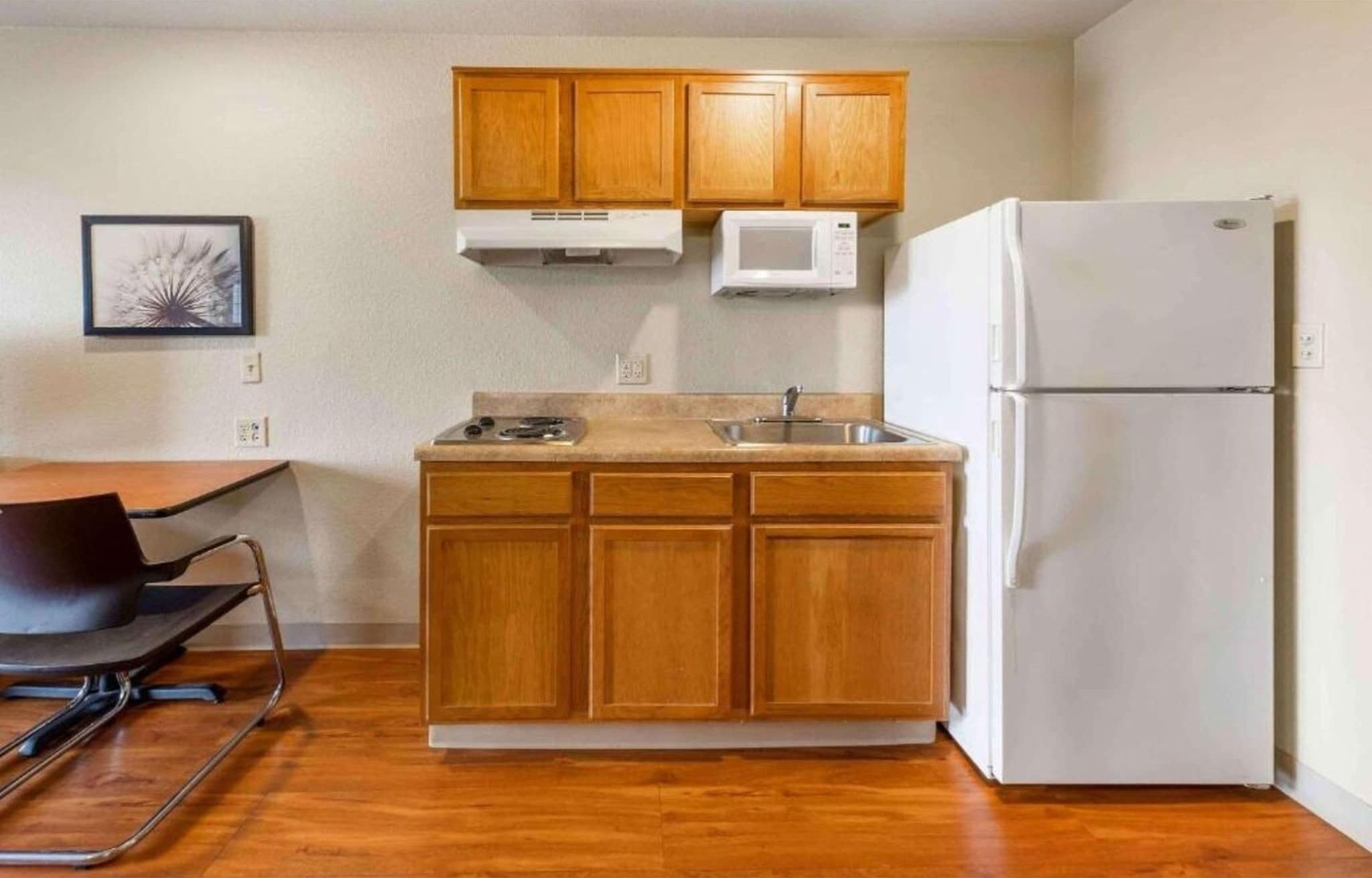 A compact kitchen with wooden cabinets, a white refrigerator, a microwave, a sink, and a chair.