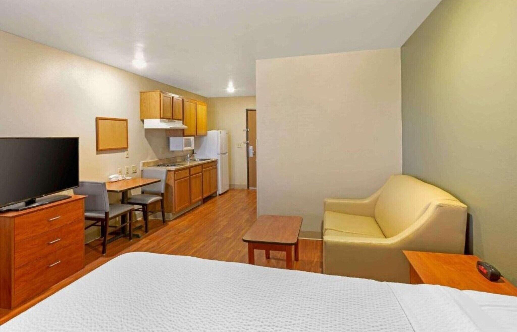 Une chambre d’hôtel de taille réduite comprenant un lit, une télévision, un petit bureau avec une chaise, une kitchenette et un canapé.