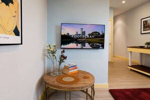 Ein modernes Interieur mit einem runden Holztisch, einem an der Wand montierten Fernseher, der 'FOOTSCRAW VICTORIA' anzeigt, und einem gerahmten Kunstwerk.