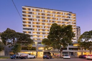 A multi-story hotel with a modern design, featuring balconies on each floor, a parking area with several cars, and a street view with trees and a building in the background. - Exclusive 1-room apartment in an elevated location, just steps away from shops. (Southbank)
