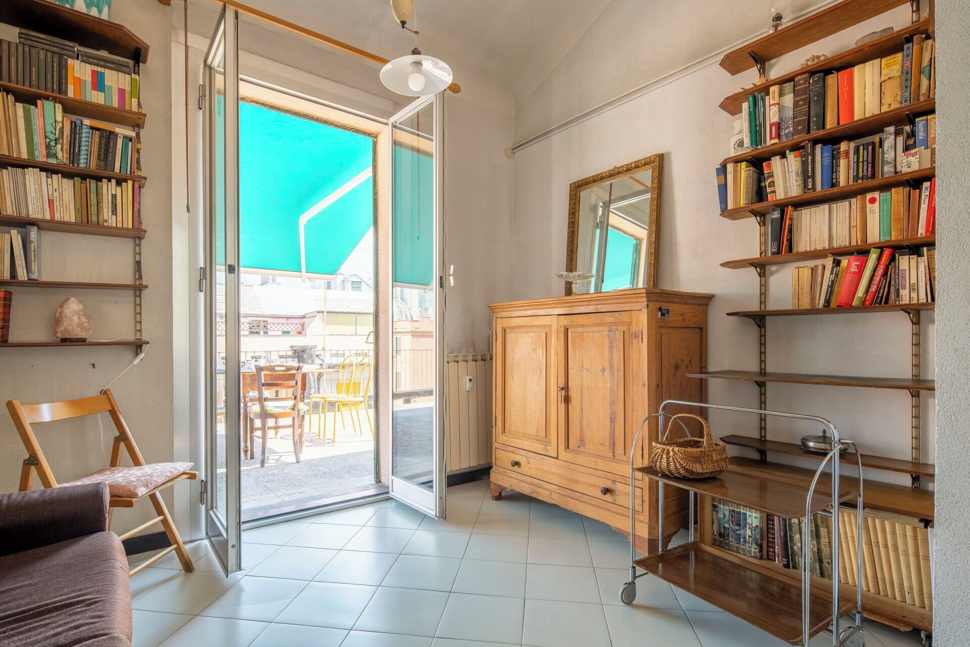 A room with a large open door leading to a balcony, a wooden cabinet, and a bookshelf filled with books.