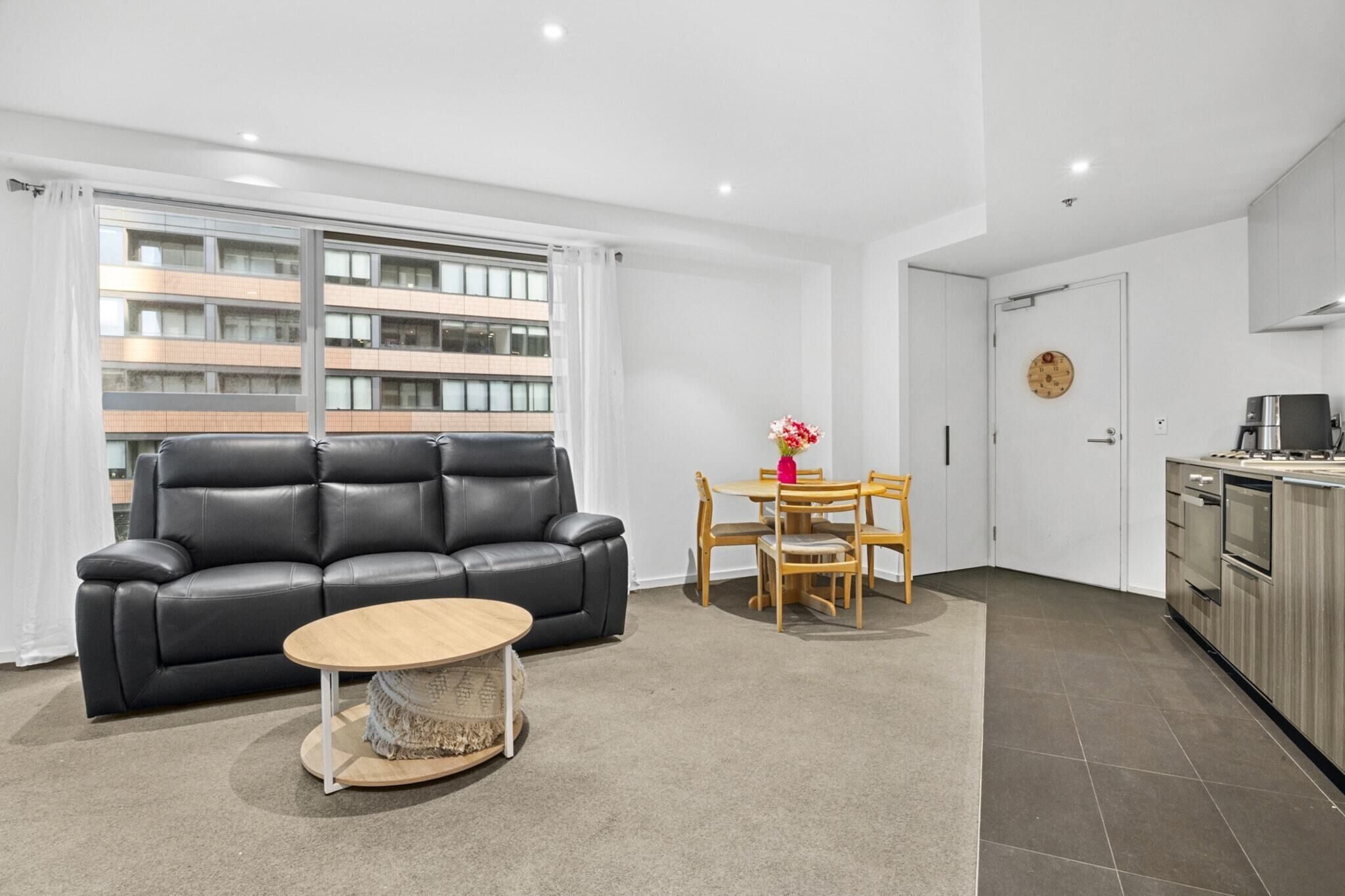 A modern living room with a black leather sofa, a round wooden coffee table, and a dining area with wooden chairs and a small table.