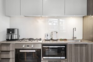 A modern kitchen with stainless steel appliances, including a coffee maker, toaster, and microwave, with wooden cabinetry and a window above the sink. - Stylish 2-Bed Apartment by 888 Collins (South Melbourne)