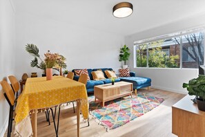 Ein modernes Wohnzimmer mit einem blauen Sofa, einem gelben Tisch und einem großen Fenster mit Blick auf Bäume.