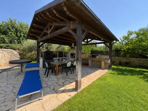 Een terras buiten met een stenen vloer, een overdekte zithoek en een barbecue.