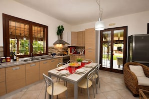A modern kitchen with a dining area, featuring a central island, wooden cabinets, and a large window with a view of greenery. - Villa in Donnalucata with 4 bedrooms sleeps 10 (Donnalucata)