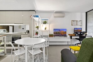 A modern kitchen with white cabinetry, a dining area with a round table and chairs, a living area with a sofa and a TV, and a cityscape painting on the wall. - City-View 2-Bed Apartment Melbourne CBD (Melbourne)
