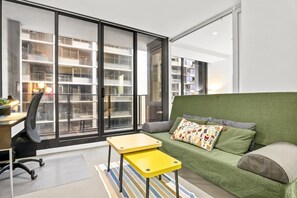Ein modernes Wohnzimmer mit einem grünen Sofa, einem gelben Beistelltisch und einem großen Fenster mit Blick auf ein mehrstöckiges Gebäude.