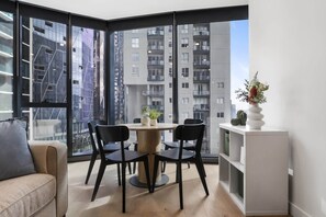 A modern living room with a round table and chairs, a sofa, a white cabinet, and a large window with a city view. - High-Rise 1-Bed Apartment Near Docklands (Melbourne)