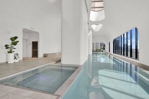 Une piscine intérieure moderne dotée d’un plancher en verre, de murs blancs et offrant une vue sur la ville.