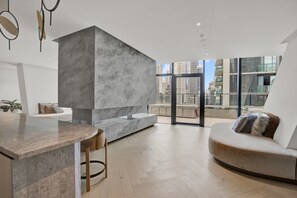 A modern interior with a kitchen area, a seating area with a sofa and cushions, and a view of the cityscape through large windows. - High-Rise 1-Bed Apartment Near Docklands (Melbourne)