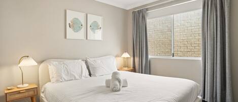 Habitación de hotel con una cama grande, dos mesitas de noche con lámparas, un ventanal con cortinas y dos cuadros en la pared.