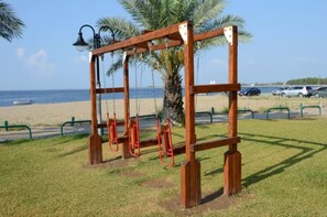A wooden swing set with chains, located on a grassy area near a beach with a palm tree and a lamp post in the background. - Halikidiki, Family house to enjoy by the beach (Kalives Polygyrou)