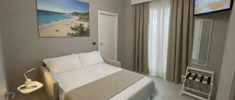 A hotel room with a bed, a television, a painting of a beach, and a window with curtains.