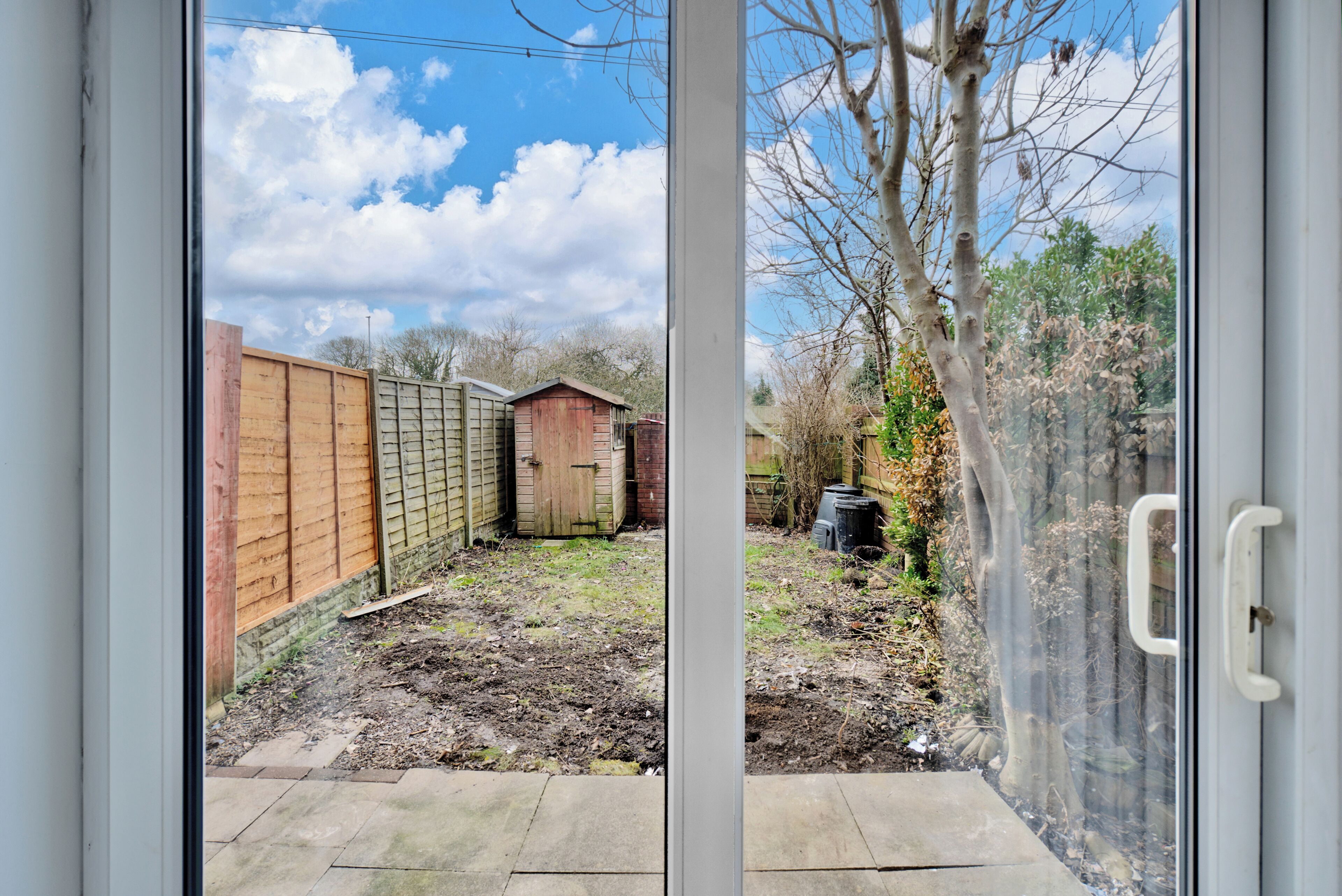 A view through a glass door of a backyard with a wooden fence, a shed, and a garden area.