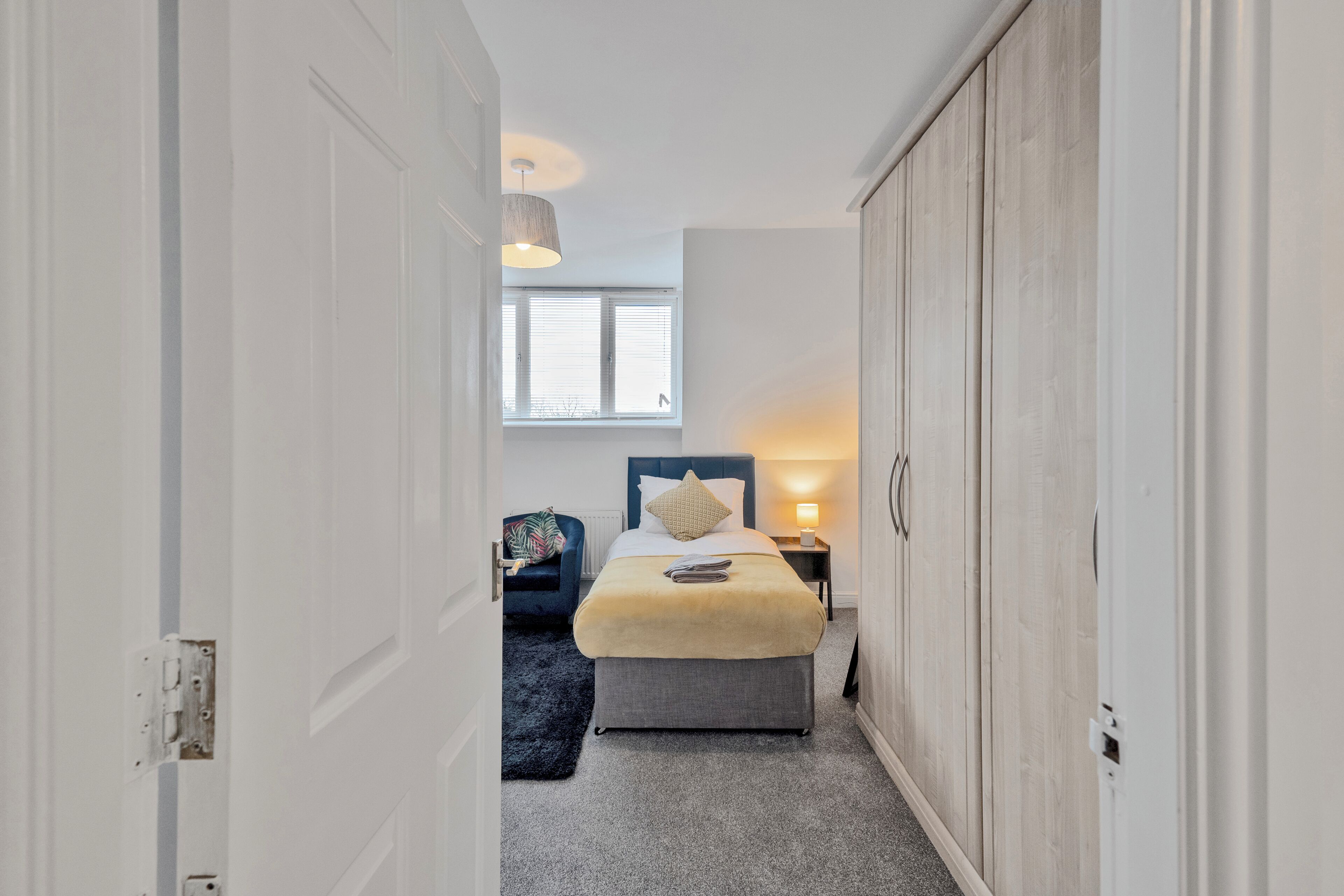 A narrow hallway leading to a bedroom with a bed, bedside table, and a window.