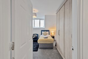 Ein schmaler Flur führt zu einem Schlafzimmer mit Bett, Nachttisch und Fenster.