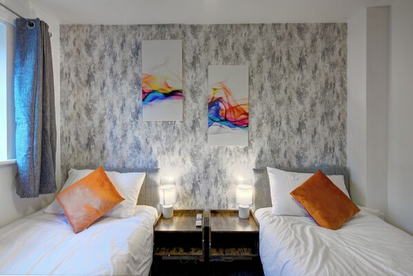 A hotel room with two beds, each with an orange pillow, a wooden nightstand with a lamp, and a textured wall with abstract paintings.