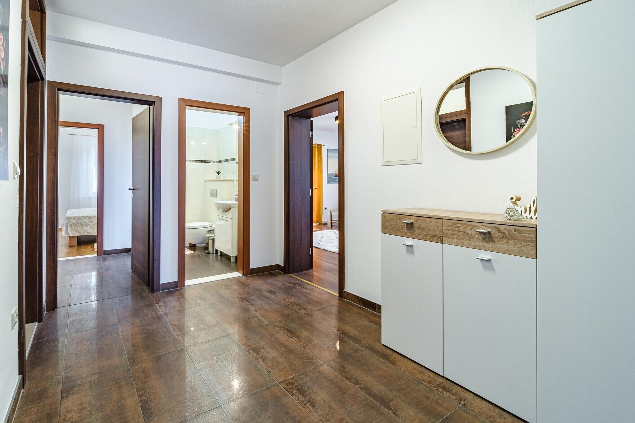 Un pasillo con piso de madera que conduce a un baño con inodoro y lavamanos. Hay puertas a ambos lados del pasillo.