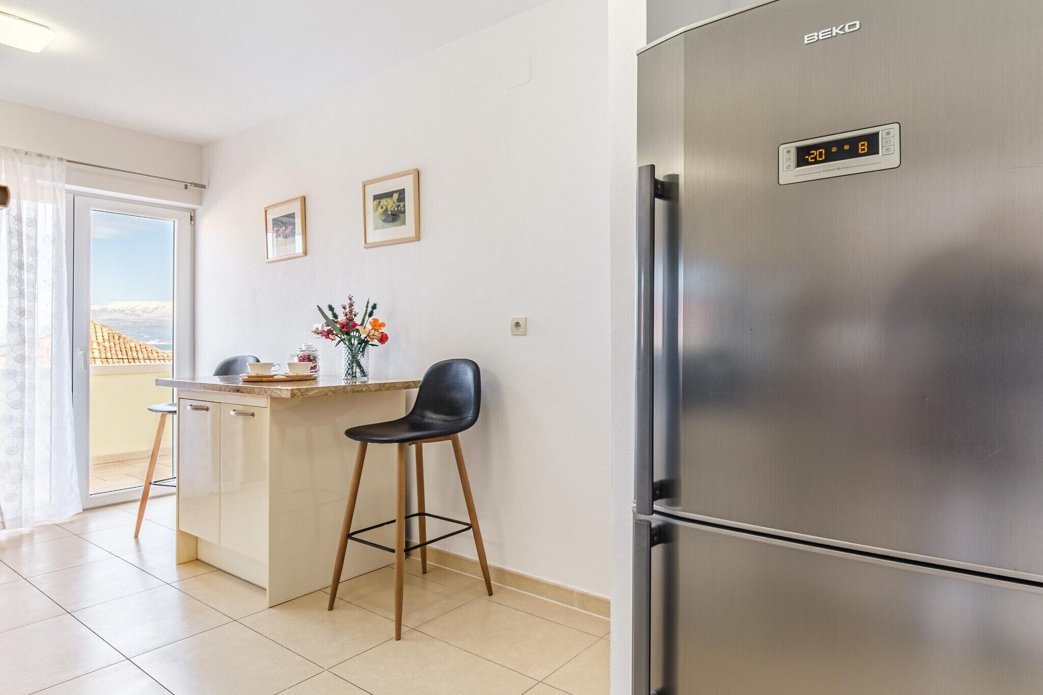 Una cocina moderna con un refrigerador de acero inoxidable, una pequeña isla con una silla y vistas a través de la puerta de cristal.