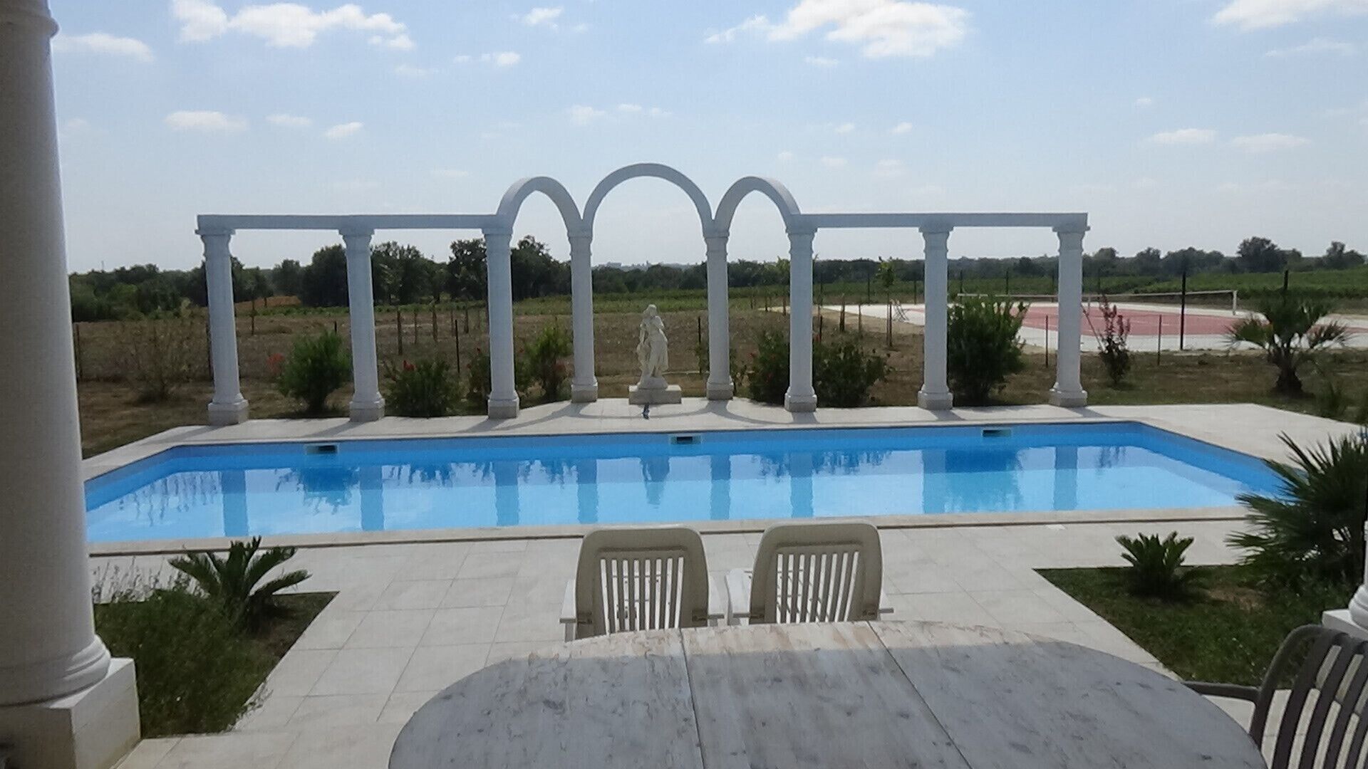 Una piscina amb una estructura de columnes blanques al fons, envoltada per una zona enjardinada.