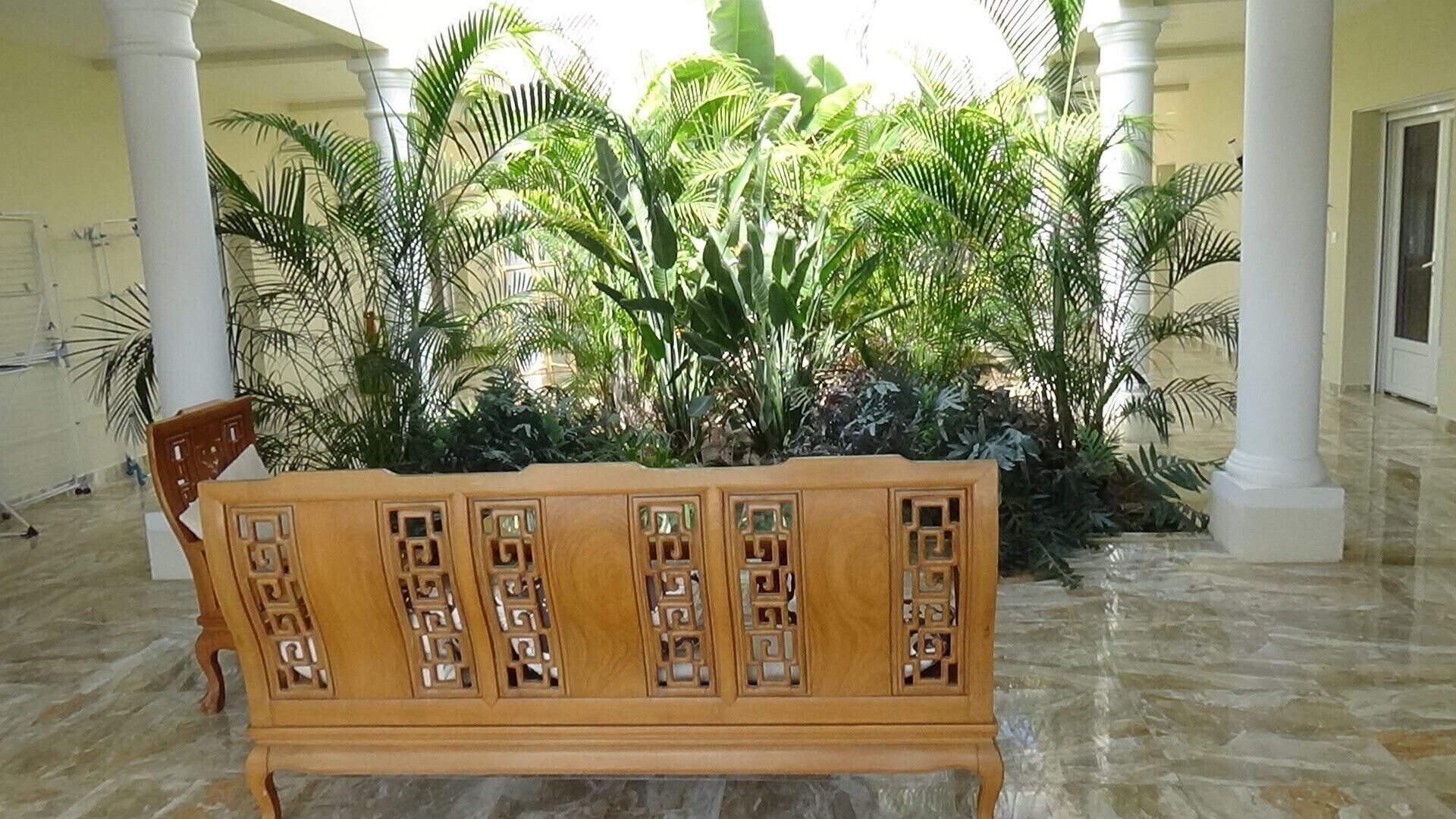 Un banco de madera con intrincados grabados, ubicado en un espacio cubierto al aire libre con piso de baldosas y exuberante vegetación tropical de fondo.