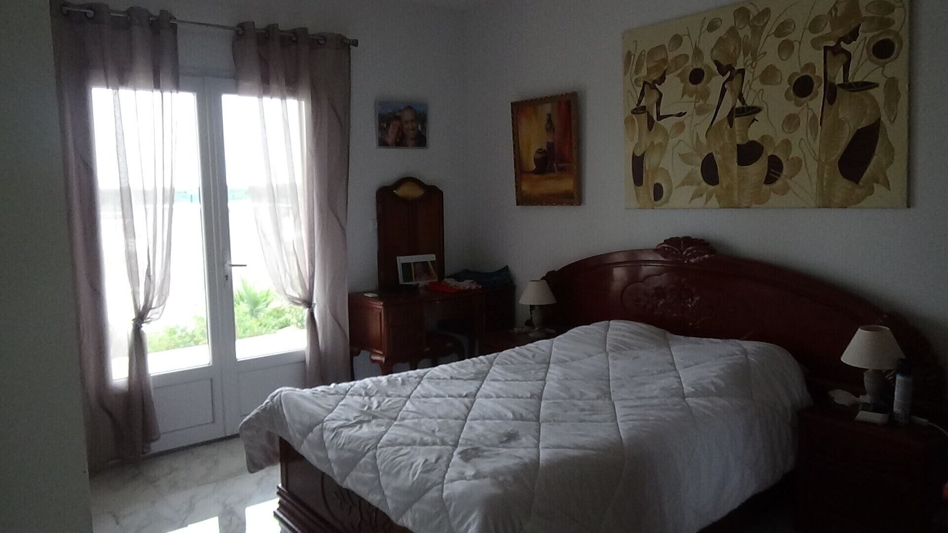 A bedroom with a bed, a nightstand, a lamp, a dresser, a painting, and a window with curtains.