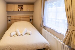 A small, single-bed room with a wooden headboard, a window with curtains, and a radiator. - Mobile Home with Shared Pool (Zonhoven)