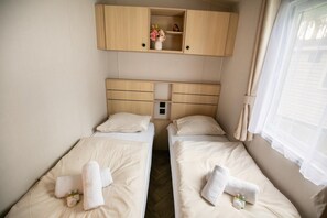 A compact room with two single beds, a small shelf with flowers, and a window with blinds.