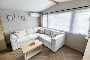 A modern living room with a sectional sofa, a wooden coffee table, and a large window with sheer curtains. - Mobile Home with Shared Pool (Zonhoven)