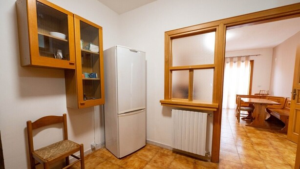 A compact kitchen with a refrigerator, wooden cabinets, a chair, and a window.